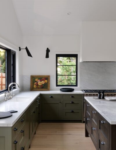 Modern kitchen with green cabinets, white countertops, black fixtures, and a black window. There is a painting on the countertop and a view of outdoor greenery through the window.