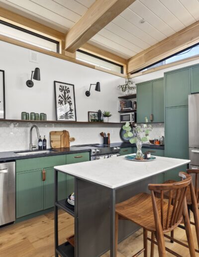 Modern kitchen with green cabinets, stainless steel appliances, a white island, and wooden stools. Shelves with decor, artwork on walls, and a wooden ceiling beam complete the space.
