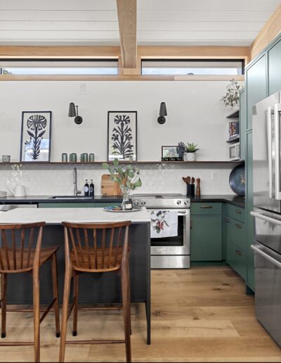 Modern kitchen with green cabinets, stainless steel appliances, and wooden beams on the ceiling. A small island with two wooden stools is central. Wall décor includes framed art and shelves with items.