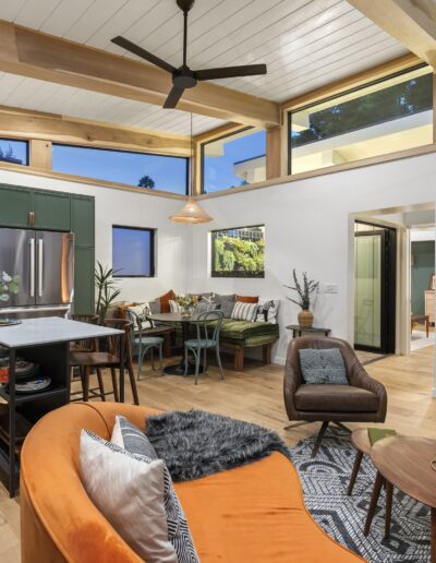 A modern open-plan living space features a kitchen with green cabinets, a dining area, and a cozy seating area with an orange couch, grey rug, and wood accents. The room includes large windows and a ceiling fan.
