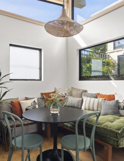 A cozy dining area featuring a round black table with a vase of flowers, surrounded by three chairs and a bench with assorted cushions, under natural light from large windows and a wicker pendant light.