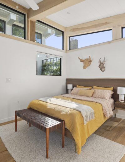 A modern bedroom with a wooden bed, yellow bedding, wicker chair, and wall-mounted deer head decor. Large windows provide natural light. An open doorway leads to an adjacent kitchen visible in the background.