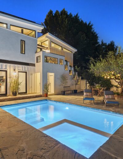 Modern two-story house with large windows and a well-lit backyard featuring a swimming pool, patio furniture, and surrounding trees, taken at dusk.