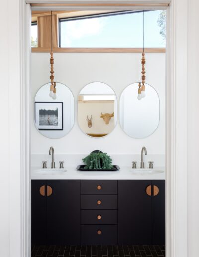 A bathroom with a double sink vanity, black cabinets, two oval mirrors, and hanging pendant lights. A potted fern sits between the sinks. A framed photo and two mounted animal heads are on the back wall.