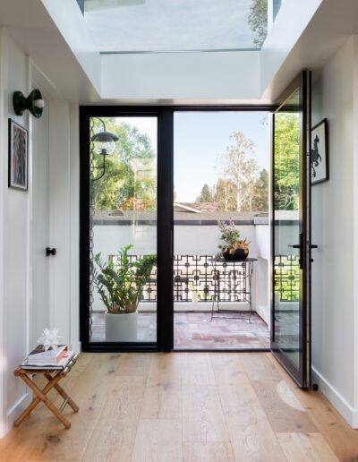 A hallway with a glass ceiling and hardwood floor leads to an open glass door, revealing a small balcony with potted plants and outdoor decor.