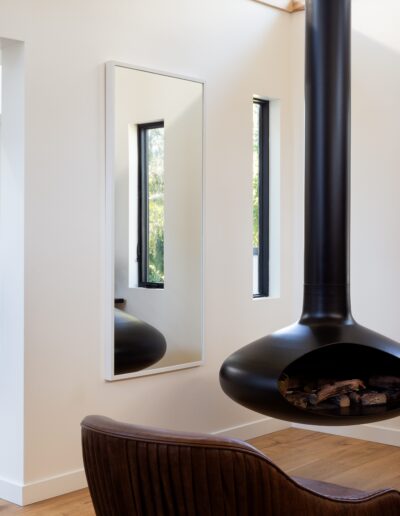 A cozy living area with a suspended black fireplace, a chair, and a large mirror. In the background, a bathroom with double sinks and round mirrors is visible.
