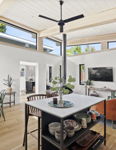 A modern, open-concept living space with large windows, a kitchen island, various indoor plants, a ceiling fan, and contemporary furniture including a green dining area and an orange armchair.