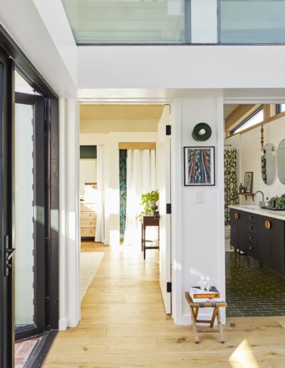 A well-lit hallway with light wooden floors leads to three rooms: an entryway with a vase of flowers, a bedroom, and a bathroom with dark cabinetry and tile flooring. A small stool with books sits in the hall.