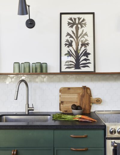 A kitchen with green cabinets, a black countertop, a stainless steel sink, and a framed plant artwork. There are glass cups on the shelf, and a cutting board with vegetables on the counter.